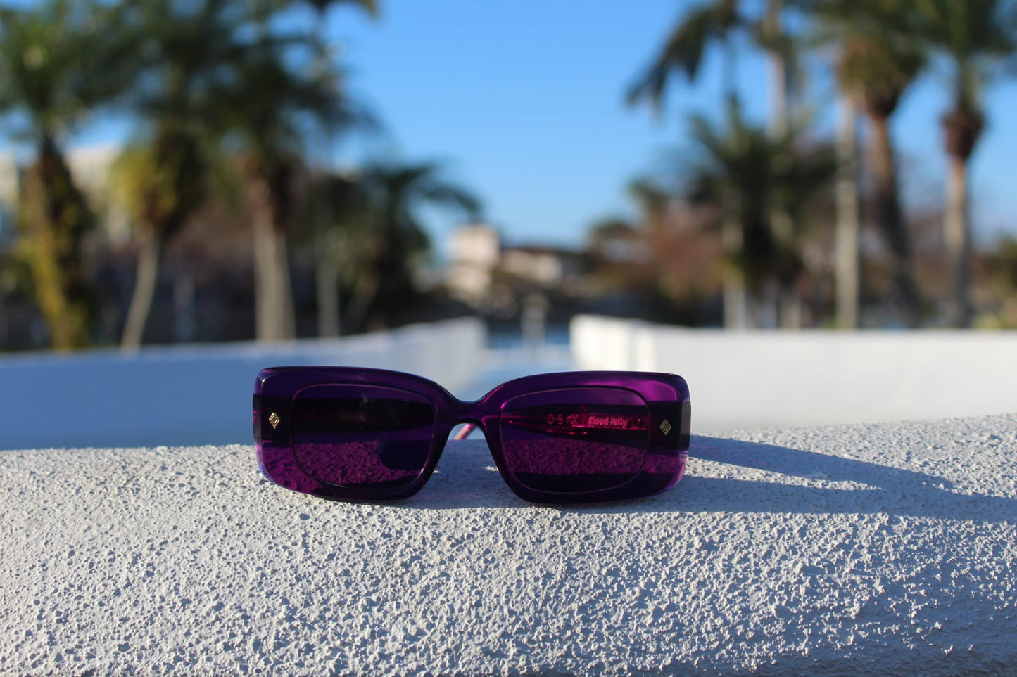 “Front view of Cloud Jelly Gift sunglasses on textured white ledge, featuring deep purple tones against a tropical city backdrop”